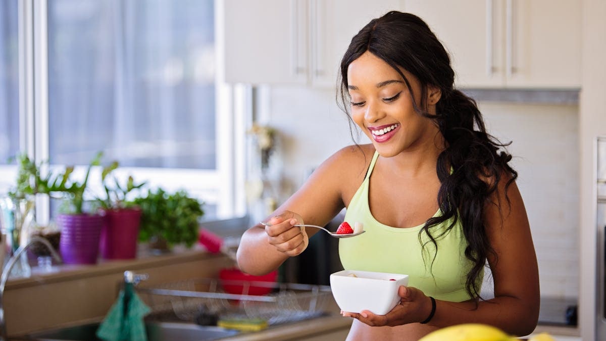 Smiling woman eating a nutritious fruit bowl in a bright kitchen setting.